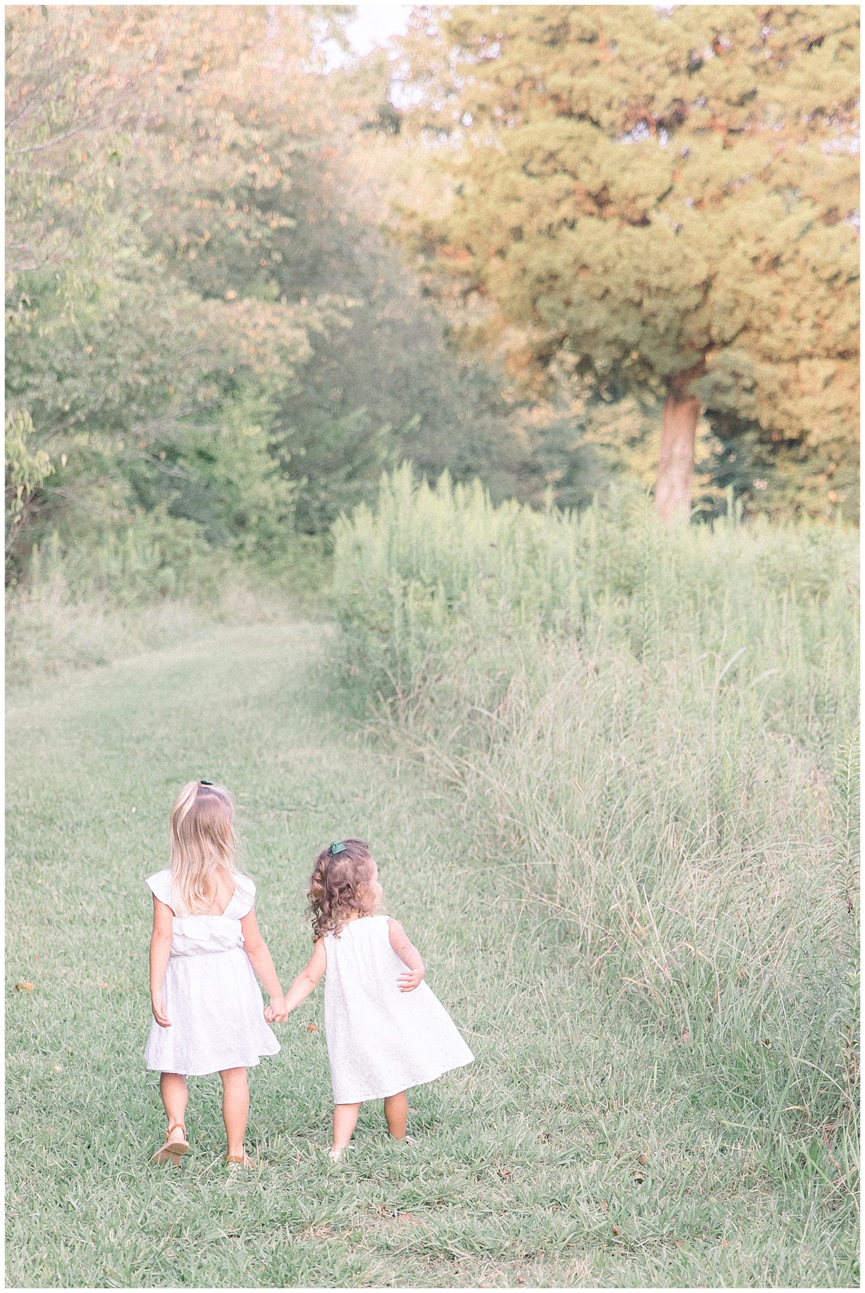 Sisters running away from camera holding hands in a tall grass field. Charlotte NC family photography